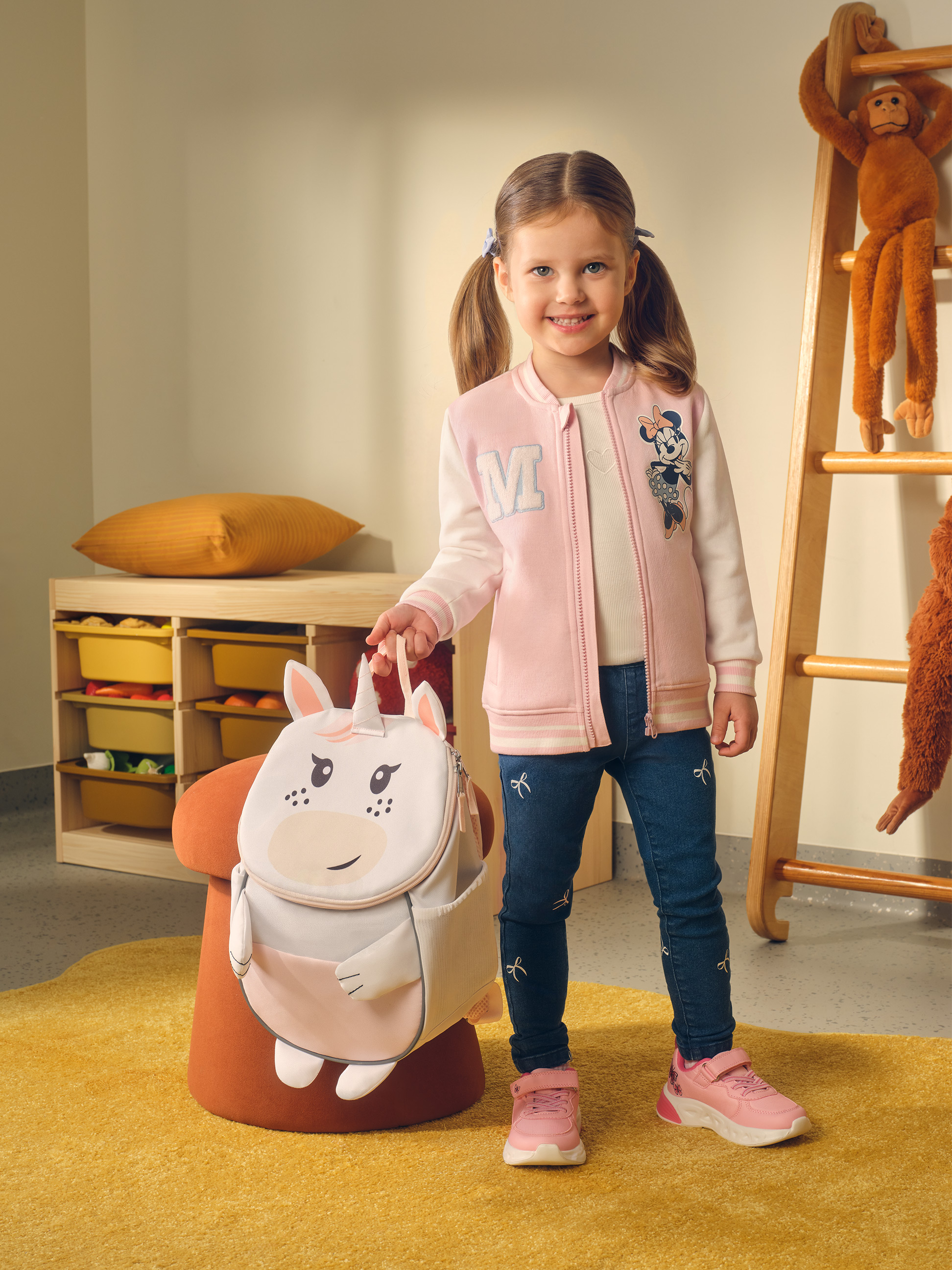 Lachend meisje met een eenhoornrugzak, roze jasje en jeans.