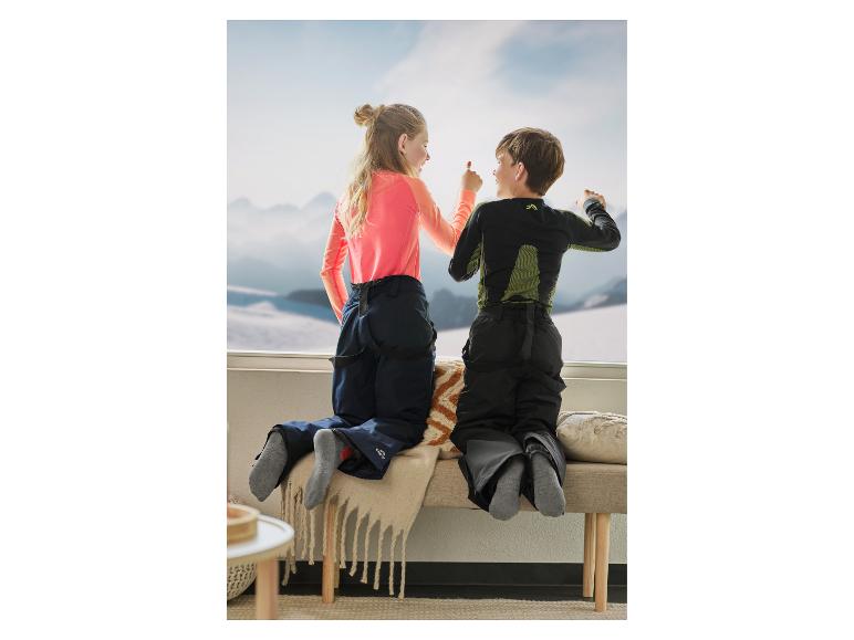 Twee kinderen in skikleding kijken uit een raam naar besneeuwde bergen.