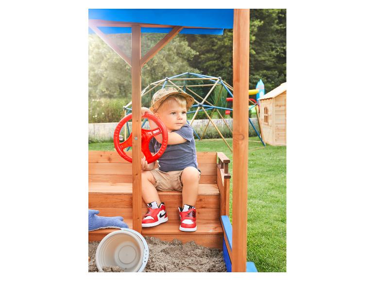 Peuter speelt in een houten zandbak met een rood stuur, draagt een hoed en sneakers.