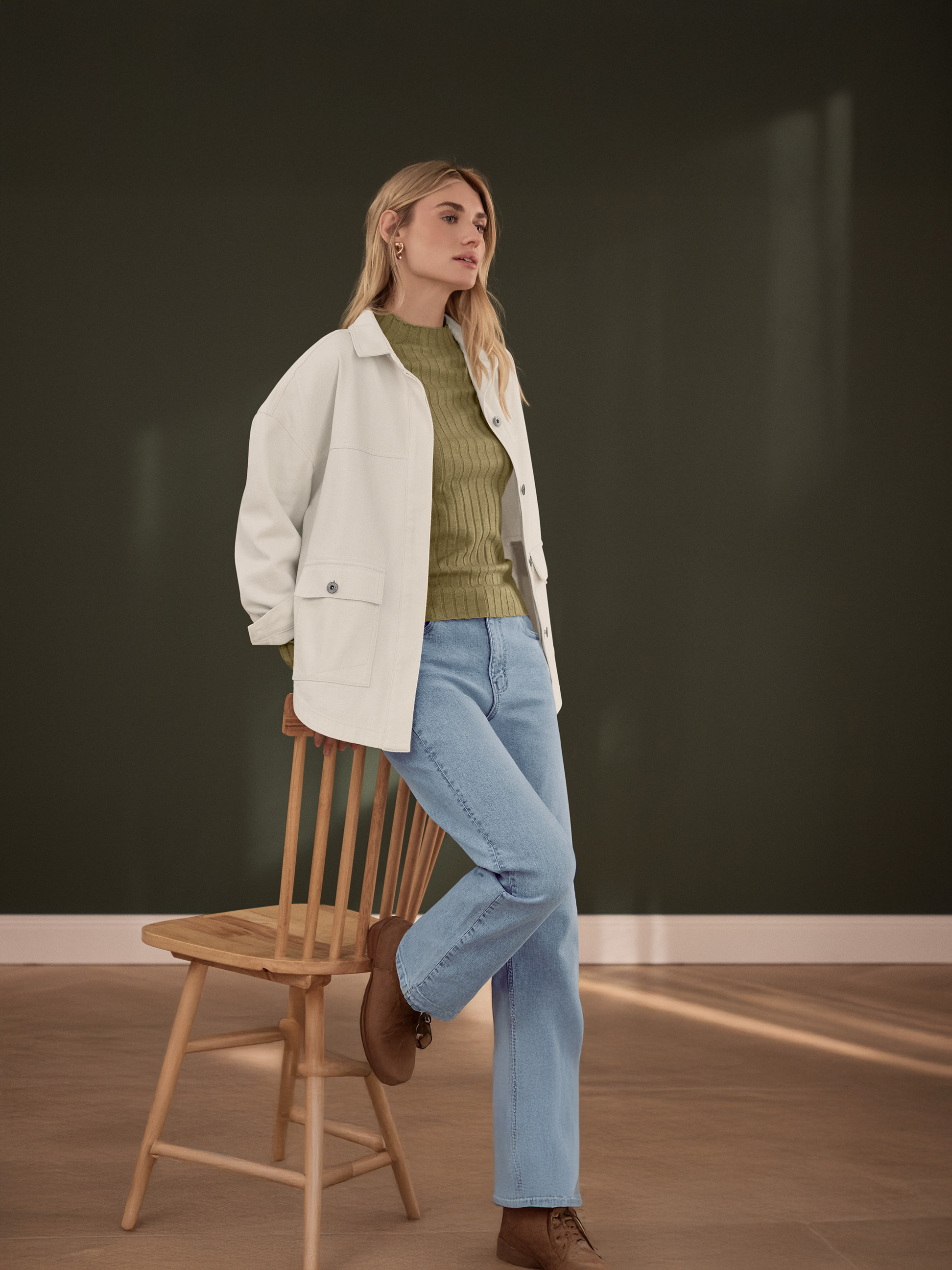 Vrouw in een wit jack, groene geribbelde top en lichtblauwe jeans, leunend op een houten stoel.