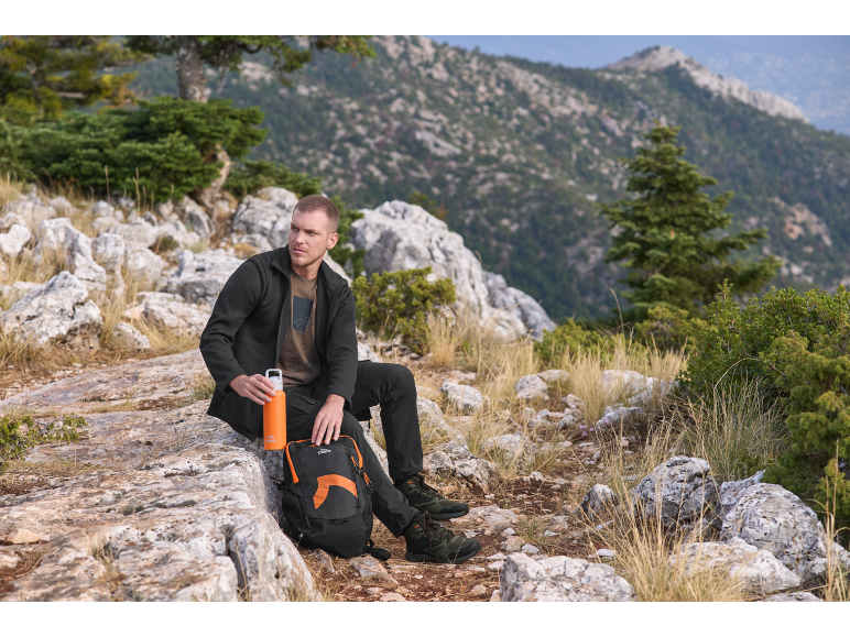 Man in fleecejack en trekkingbroek zit op een rots met rugzak en fles.