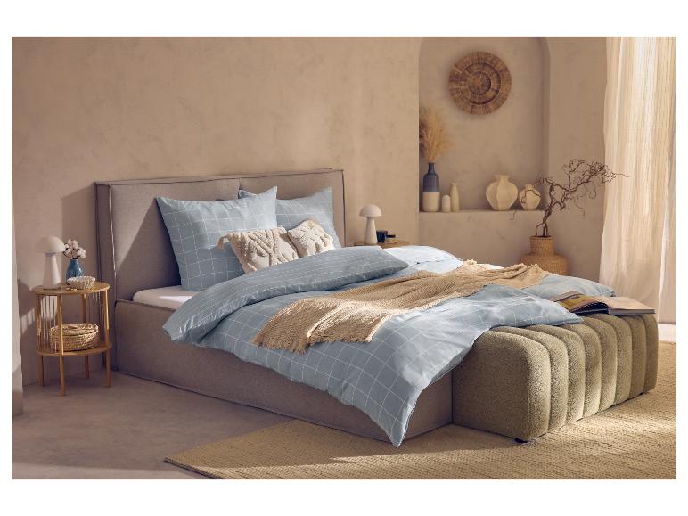 Slaapkamer met lichtblauw geruit beddengoed, lichtbruin bedframe en poef, licht tapijt.