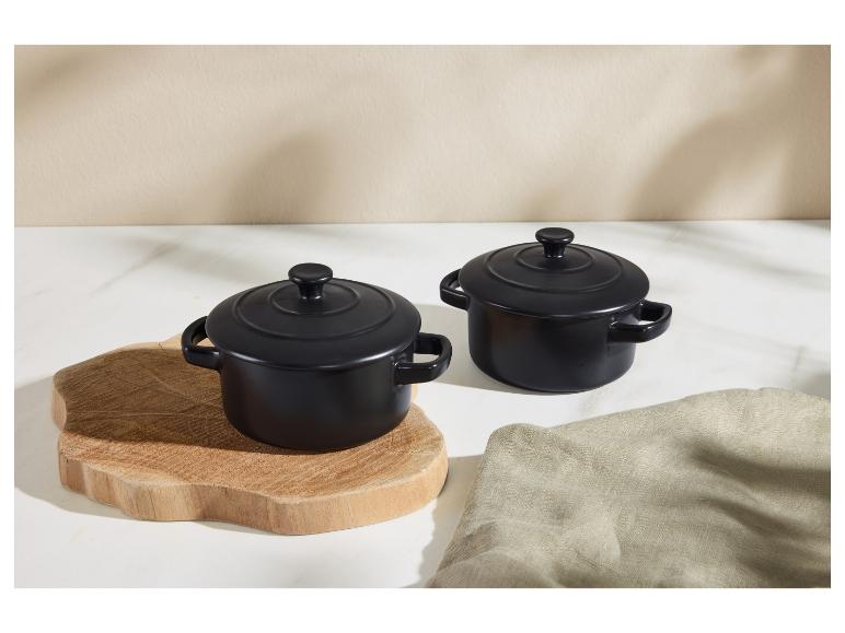 Twee zwarte keramische mini-cocottes met deksels, één op een houten plank, op een wit oppervlak.