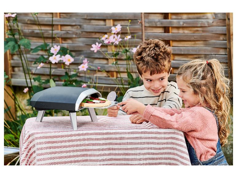 Twee kinderen bereiden speelgoedpizza in een buitenoven op een gestreepte tafelkleed.