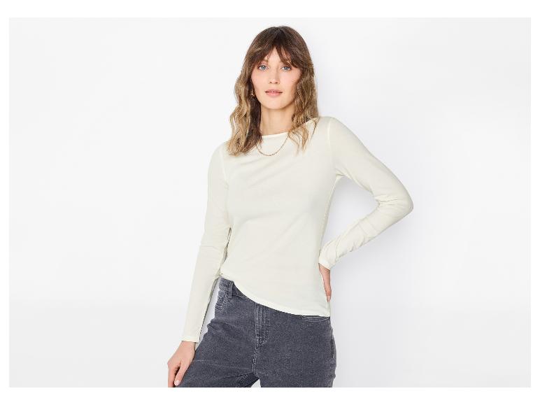 Vrouw in een lichtgekleurde blouse met lange mouwen en donkere jeans, poserend tegen een witte achtergrond.