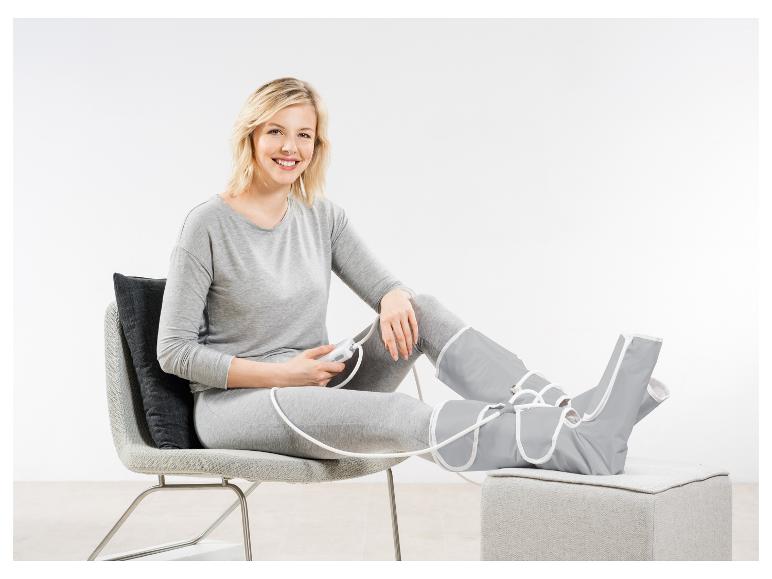 Vrouw zit op een stoel met beenmassageapparaat, glimlachend.