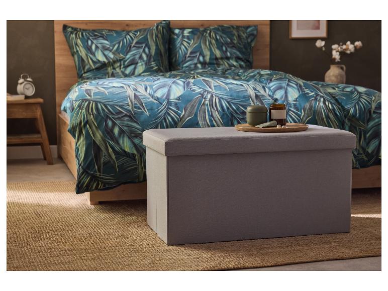 Een grijze opbergpoef van stof staat op een jutekleed in een slaapkamer met een bed in tropische stijl.