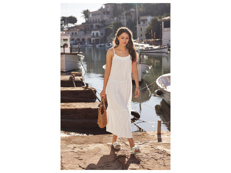 Een vrouw in een witte jurk en rieten tas, die op een kade loopt