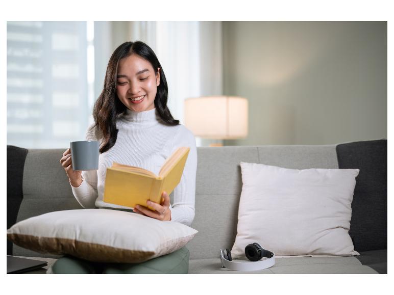 Lachende vrouw leest een boek en houdt een mok vast op een bank.