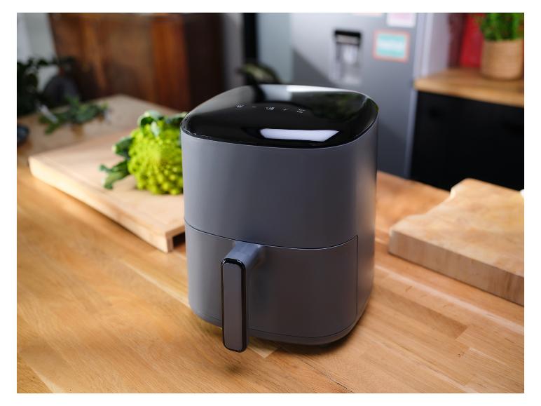 Een grijze airfryer op een houten aanrecht, met groenten op de achtergrond.