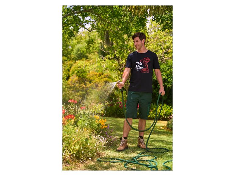 Man in zwart T-shirt en groene korte broek besproeit bloemen in een tuin.