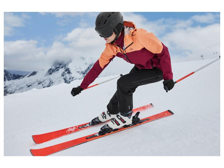 Een vrouw skiet van een besneeuwde helling in een oranje-bordeauxrode jas en zwarte broek.