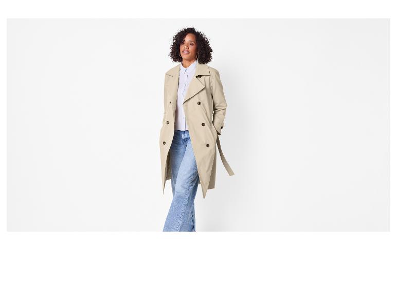 Vrouw in beige trenchcoat, lichtblauwe blouse en jeans.