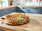 Een quiche in een glazen bakvorm op een houten snijplank in een keuken.