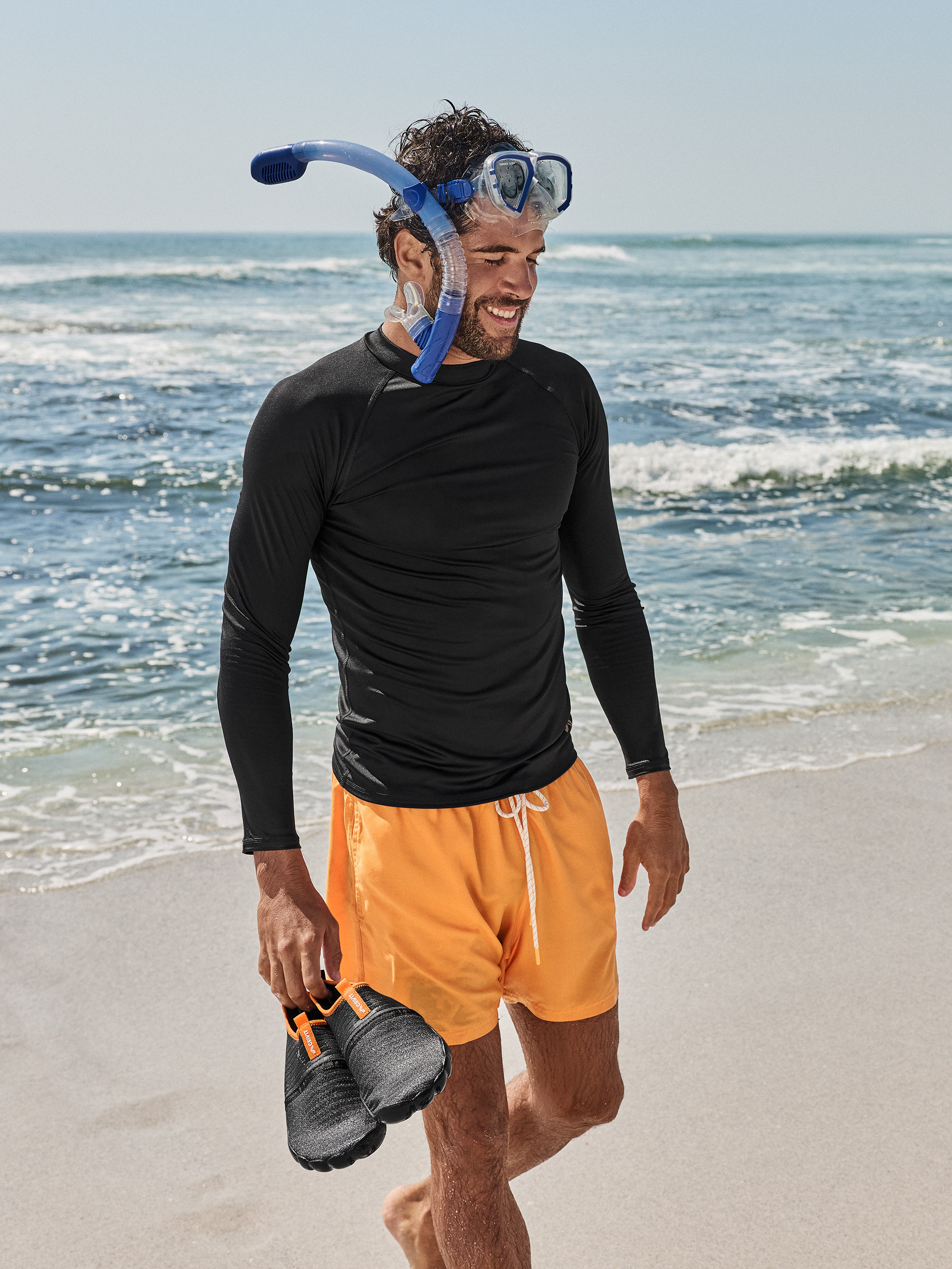 Man in zwart shirt met lange mouwen, oranje zwemshort, met duikbril en snorkel, waterschoenen vasthoudend op het strand.
