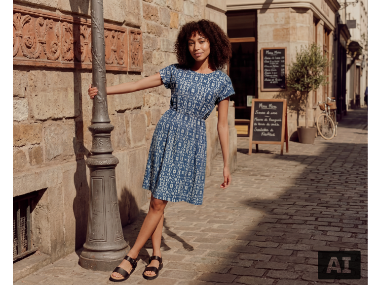 Vrouw in een blauwe jurk met patroon en zwarte sandalen in een kasseienstraat.