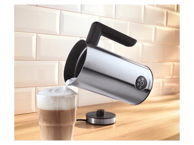 Melkopschuimer schenkt opgeschuimde melk in een koffieglas op een houten aanrecht.