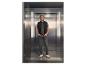 Man in een vest in een lift, draagt jeans en een t-shirt.