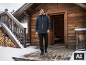 Man in donkerblauwe winterjas en zwarte broek voor een houten hut in besneeuwd landschap.