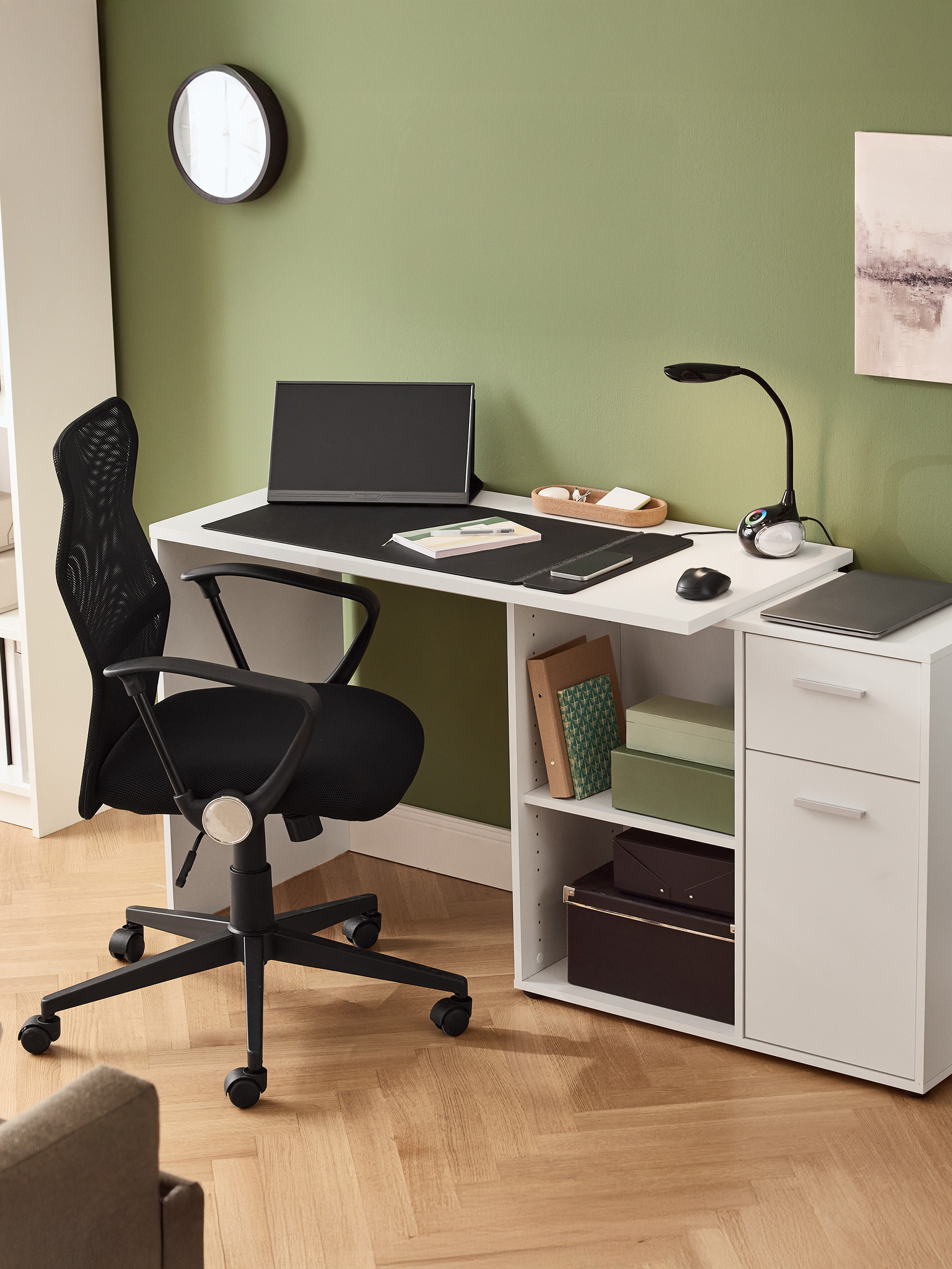 Wit bureau met zwarte bureaustoel, monitor, lamp en lades in een groene kamer.