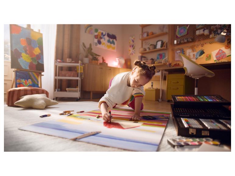 Een meisje tekent op de vloer met een grote kunstset in een kamer vol kunst.