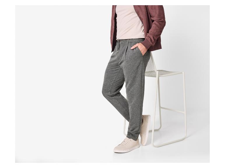 Man in grijze joggingbroek, beige t-shirt en bruin jack, leunend op een kruk.