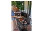 Vrouw grilt vlees en groenten op een gasbarbecue, met familie aan tafel op het terras op de achtergrond.