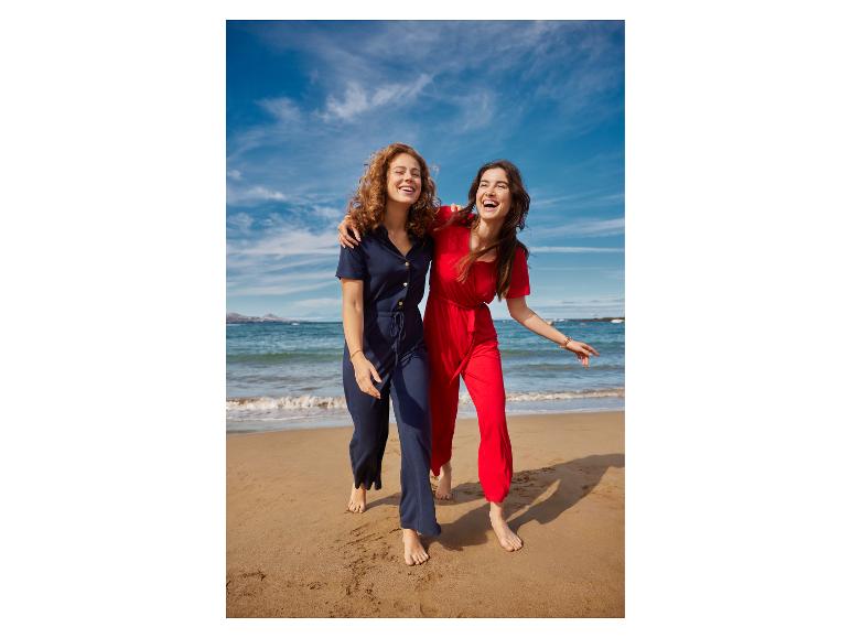 Twee vrouwen in jumpsuits op het strand, lachend en omhelzend.