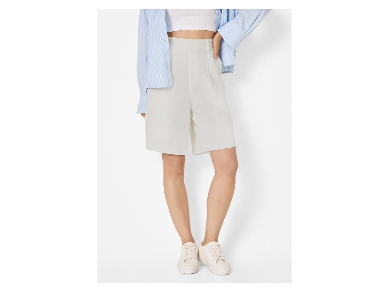 Vrouw in lichtgrijze shorts, witte crop top, blauw gestreept overhemd en witte sneakers.