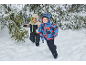 Twee jongens in winteroveralls en jassen rennen in de sneeuw naast dennenbomen.