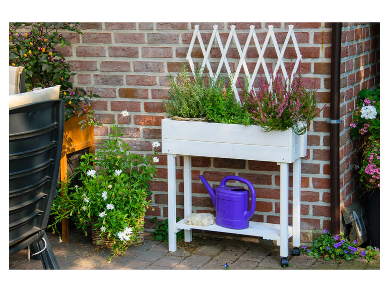 Een wit plantenrek met verschillende bloemen, een gieter en een trellis.