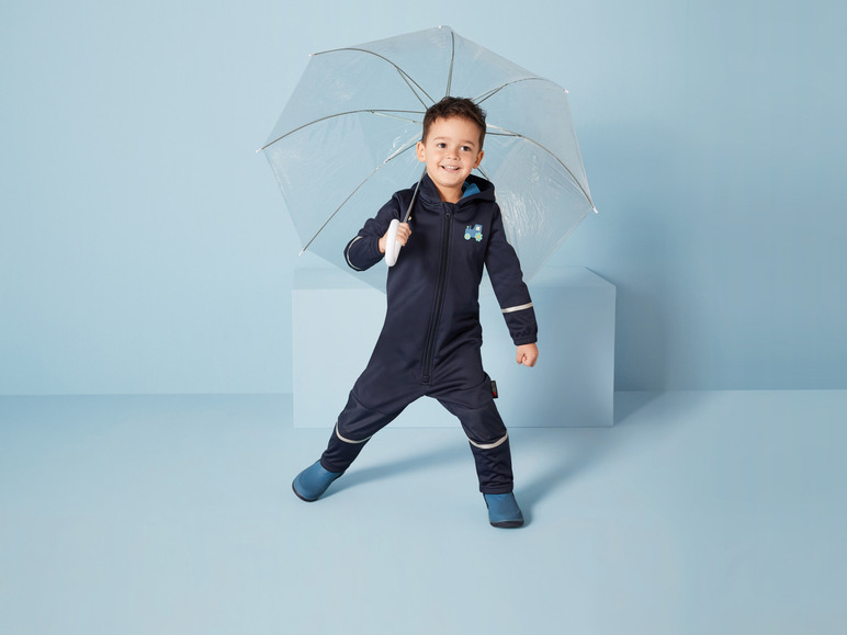 Een jongen in een blauwe regenpak met een tractorprint, met een doorzichtige paraplu.