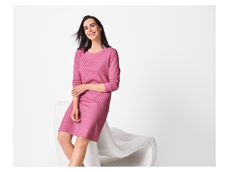 Vrouw in een roze jurk met stippen zit op een witte stoel, glimlachend.