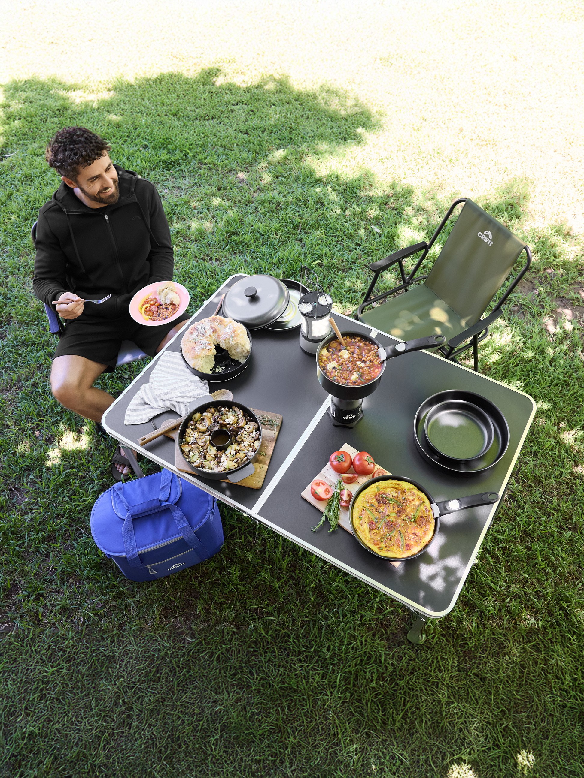 Man geniet van een maaltijd aan een campingtafel met diverse gerechten en kookgerei.