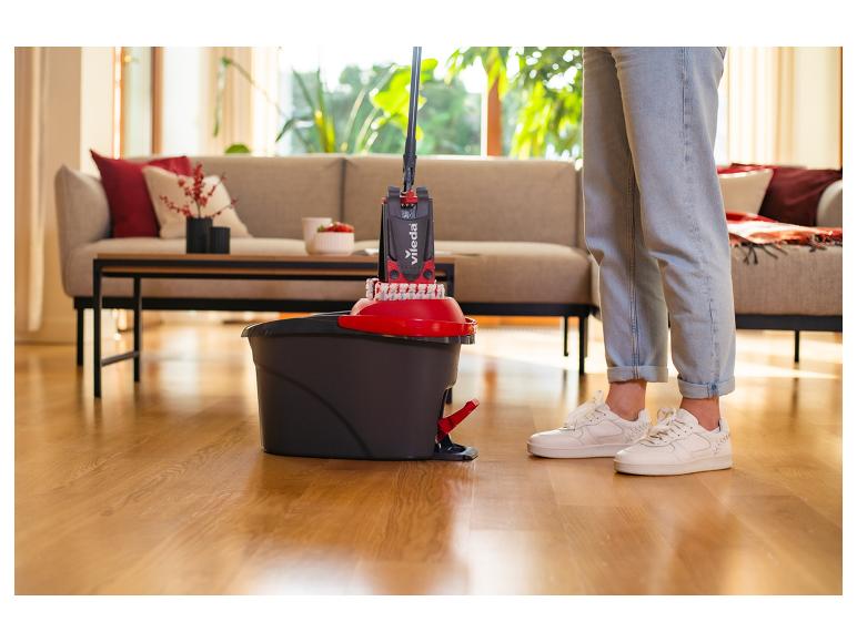 Persoon die een Vileda dweil en emmer gebruikt om een houten vloer in een woonkamer schoon te maken.