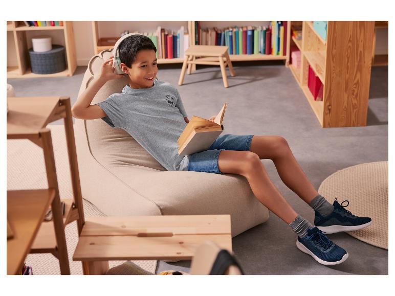 Een kind leest een boek op een fauteuil, draagt een koptelefoon en sneakers.