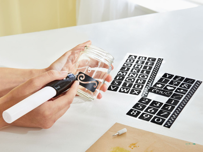 Een persoon gebruikt een sjabloon om een glazen pot te versieren met een pen.