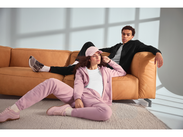 Twee jonge mensen ontspannen op een bank, gekleed in hoodies en joggingbroeken.