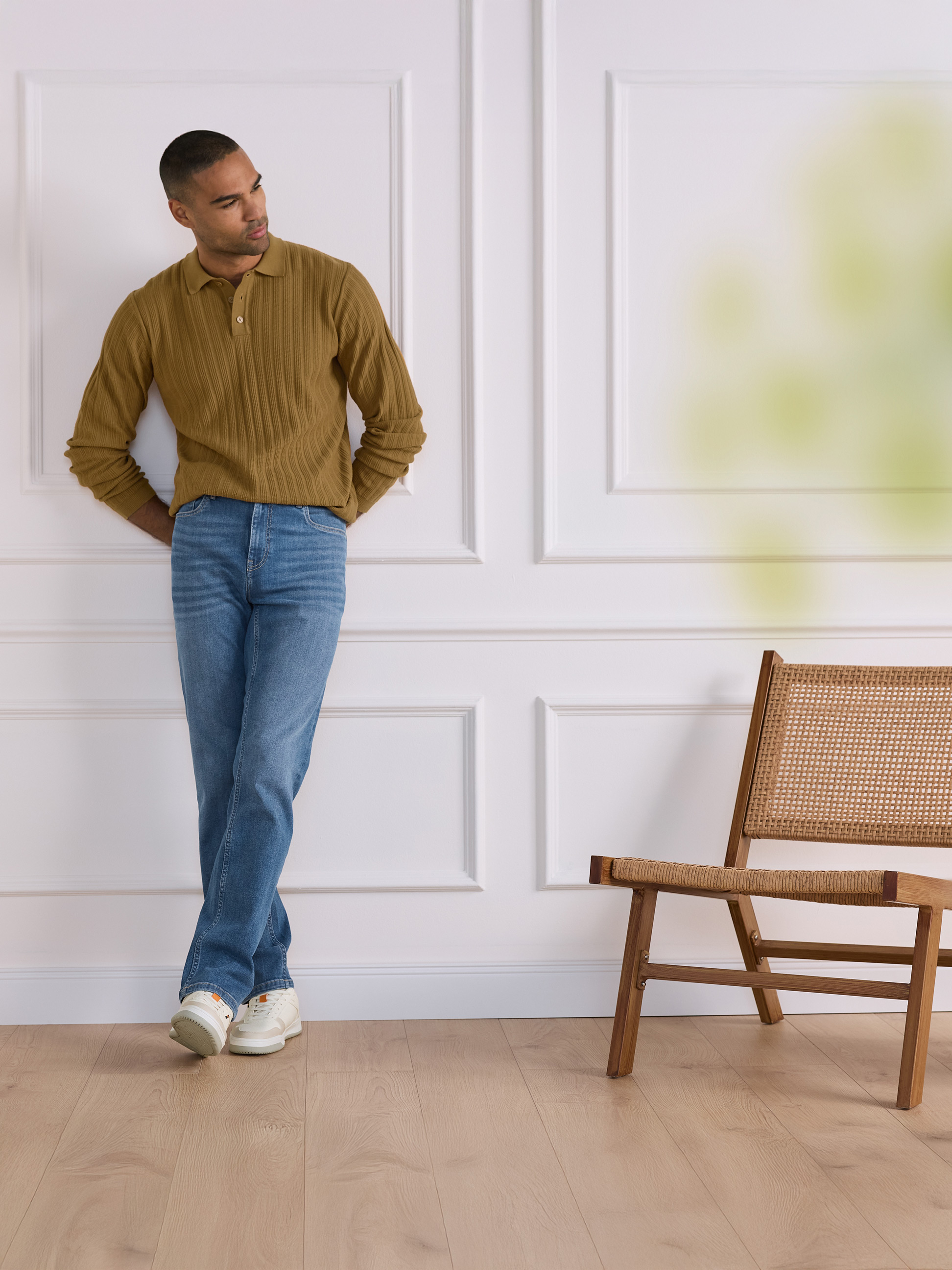 Man in bruine geribbelde polo en jeans leunt tegen een witte muur met een houten stoel.