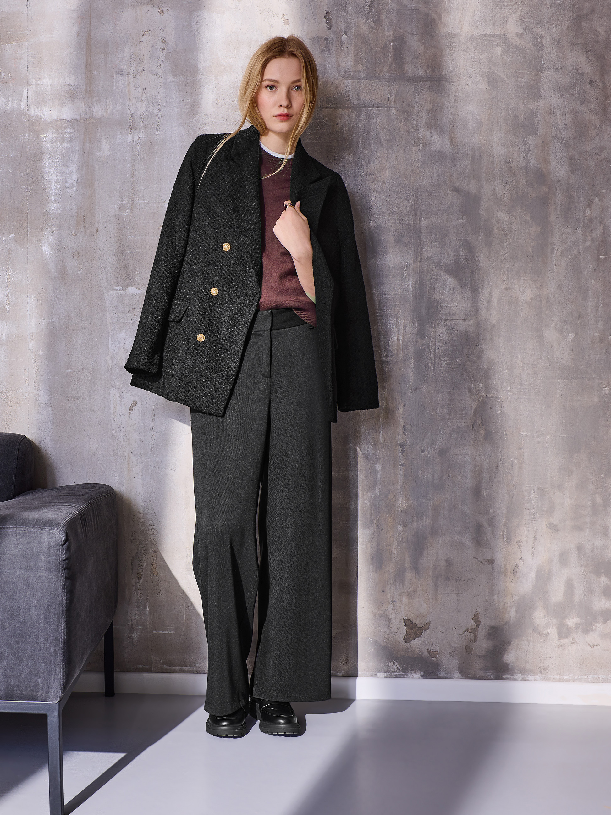 Vrouw in zwarte blazer, bordeauxrode trui en zwarte wijde broek, poserend voor een getextureerde muur.