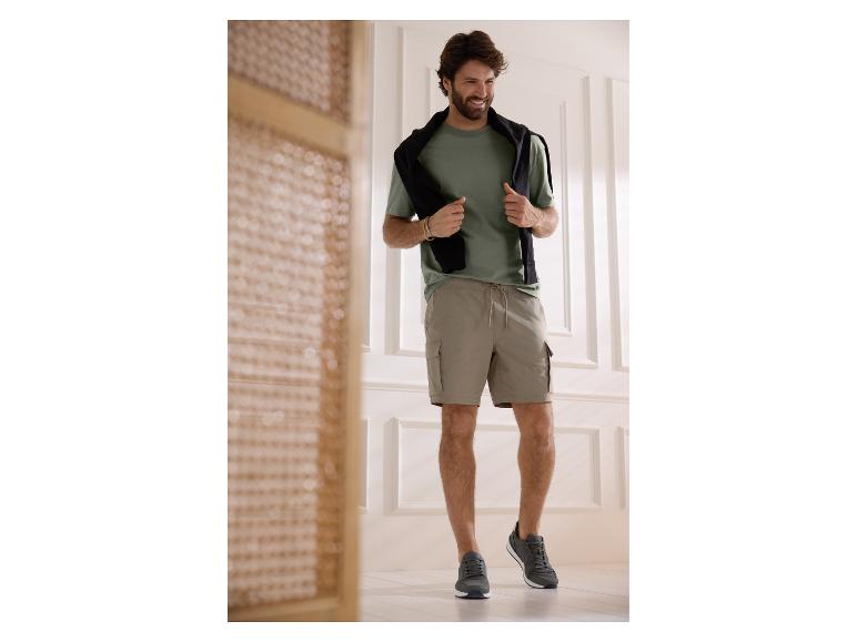 Man in groen T-shirt, beige cargoshort en grijze sneakers, lachend binnenshuis.