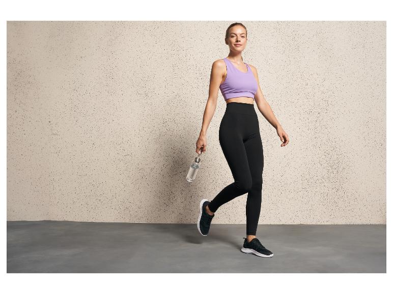 Een vrouw in zwarte leggings en een lila sportbeha poseert met een waterfles.