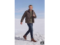 Man in bruine pufferjas, groene trui en jeans, lopend door de sneeuw.