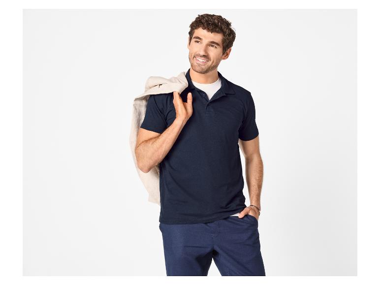 Man in donkerblauwe polo en blauwe broek, met een beige jas over zijn schouder.