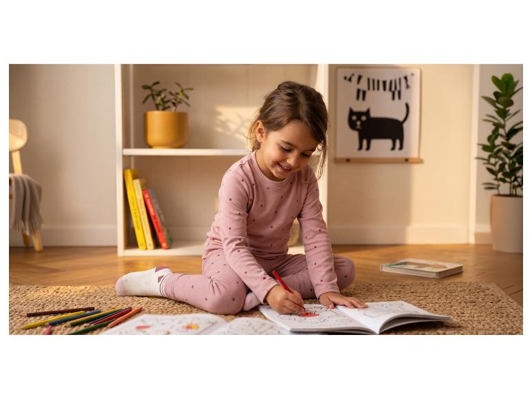 Lachend meisje in roze pyjama kleurt een boek op een vloerkleed met potloden.