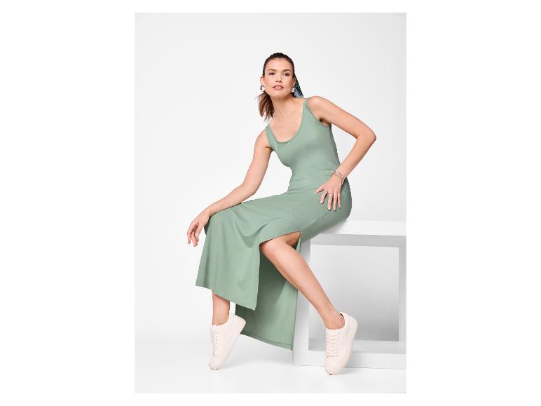 Vrouw in een groene maxi-jurk en witte sneakers, zittend op een wit voetstuk.