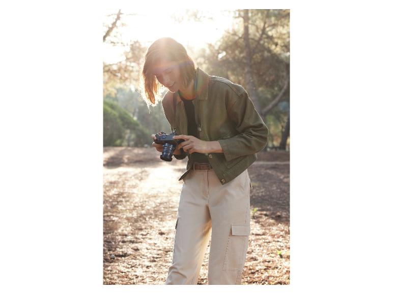 Een vrouw met een groene jas houdt een Canon camera vast in een bos.