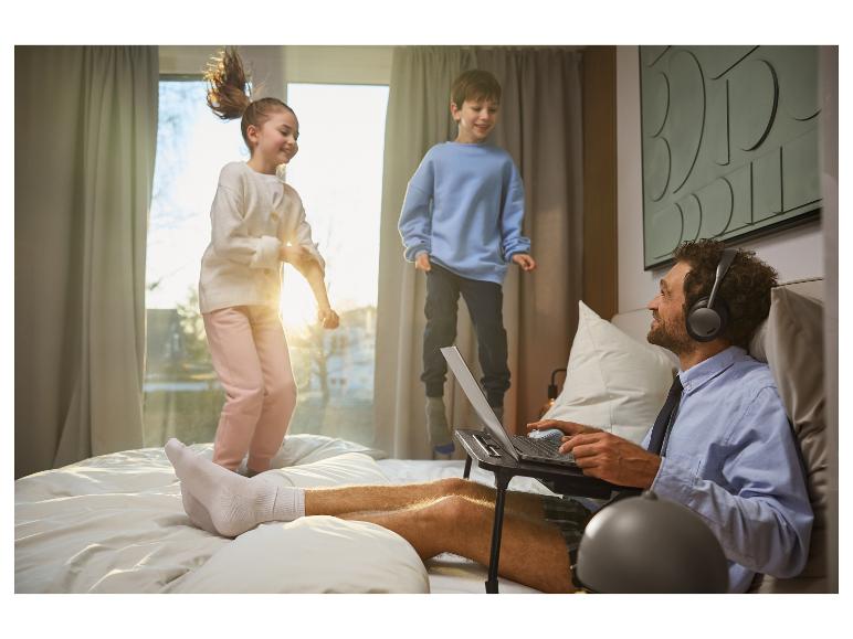 Een man werkt op een laptop in bed, draagt een koptelefoon, terwijl zijn kinderen speels op het bed springen.
