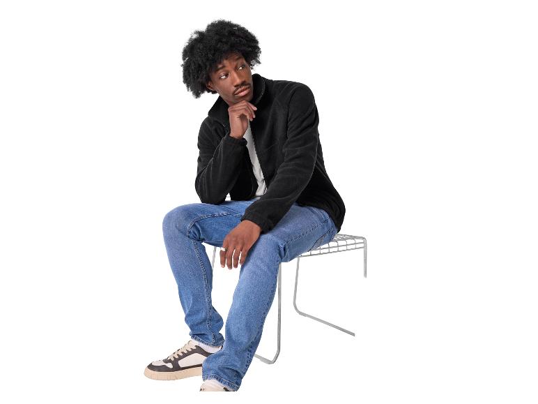 Een man in een zwart fleecejack en blauwe jeans zit op een metalen kruk, peinzend.