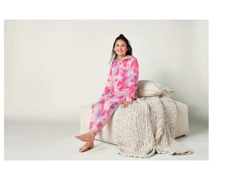 Een meisje draagt een roze tie-dye onesie en zit op een witte bank met een gebreide deken.